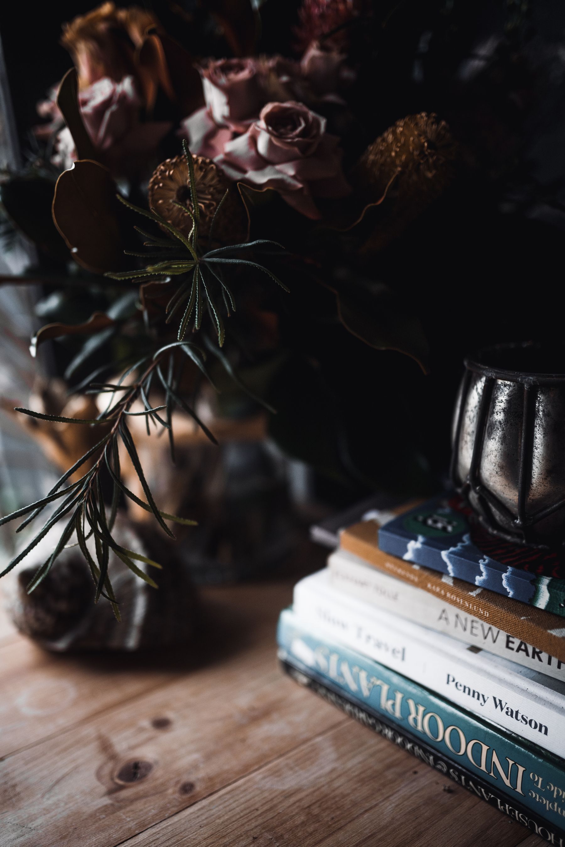 A moody still life of dusty-rose blooms, stacked books and a brass vessel in warm light